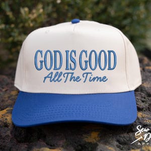 K&ouml;nnte beinhalten: Eine cremefarbene Baseballkappe mit blauem Schirm und einem blauen Knopf an der Oberseite. Auf der Vorderseite befindet sich ein gestickter blauer Schriftzug GOD IS GOOD in fetter Schrift, darunter die Worte All The Time in Schreibschrift.
