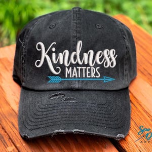 May include: A black distressed baseball cap with a white embroidered message that reads "Kindness Matters" and a blue arrow pointing to the right.
