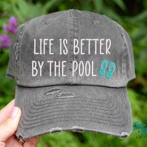 May include: A gray distressed baseball cap with the text "Life is better by the pool" in white embroidery and turquoise flip-flop icons.