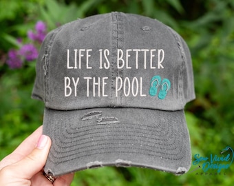 Op de afbeelding: Een grijze, verweerde baseballpet met de tekst "Life is better by the pool" in witte borduurwerk en turquoise flip-flop iconen.