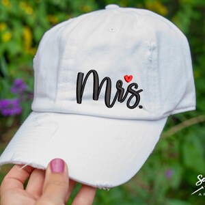 K&ouml;nnte beinhalten: Wei&szlig;e Baseballkappe mit dem in Schwarz gestickten Wort "Mrs." und einem kleinen roten Herzen &uuml;ber dem "s".