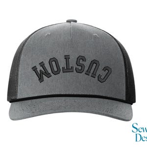 May include: Gray trucker hat with the word "CUSTOM" embroidered across the front in a dark gray, block-style font. The hat has a mesh back and a curved brim. The hat is a custom design.