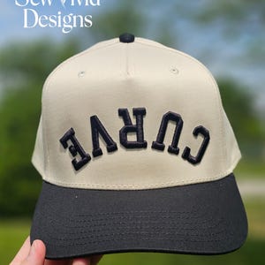 May include: A baseball cap with a cream-coloured crown and a black brim. The word "CURVE" is embroidered in navy blue across the front. The cap has a black button on top. The text "Sew Vivid Designs" is in the upper left corner.