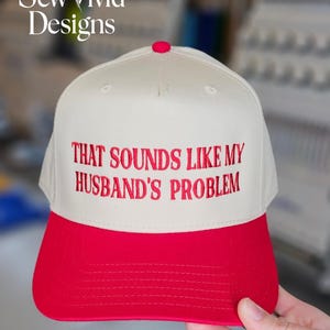 May include: A cream and red baseball cap with the embroidered text "THAT SOUNDS LIKE MY HUSBAND'S PROBLEM." The cap has a red peak and button, and the text is in red. The brand "Sew Vivid Designs" is in the upper left corner.