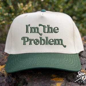 May include: A baseball cap with a cream-colored crown and a dark green brim. The phrase "I'm the Problem" is embroidered in dark green across the front. The cap has a curved brim and a button on top.