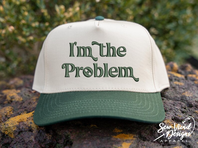 May include: A baseball cap with a cream-colored crown and a dark green brim. The phrase "I'm the Problem" is embroidered in dark green across the front. The cap has a curved brim and a button on top.
