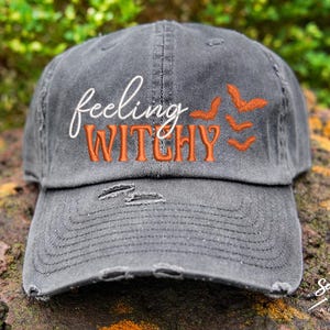 May include: A gray distressed baseball cap with the text "feeling witchy" embroidered in white and orange. The cap has a small orange embroidered bat design on the side.