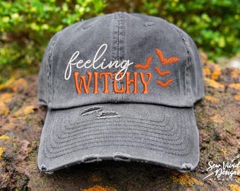 May include: A gray distressed baseball cap with the text "feeling witchy" embroidered in white and orange. The cap has a small orange embroidered bat design on the side.