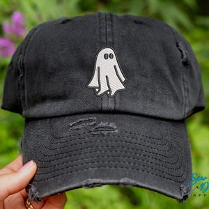 May include: A black distressed baseball cap with a white ghost embroidered on the front. The cap has a curved brim and a distressed look.
