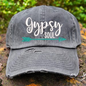 May include: A black distressed baseball cap with the text "Gypsy Soul" embroidered in white with a teal arrow pointing to the right.