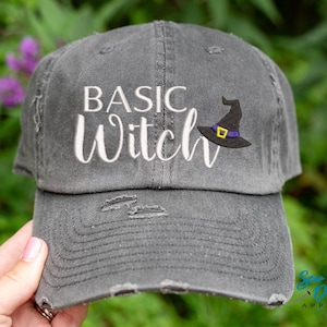 May include: A grey distressed baseball cap with the text "Basic Witch" embroidered in white and a black witch hat with a purple band embroidered on the front.