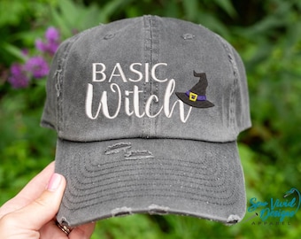 May include: A gray distressed baseball cap with the text "Basic Witch" embroidered in white and a black witch hat with a purple band embroidered on the front.