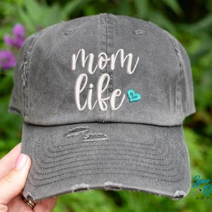 May include: A grey distressed baseball cap with the text "mom life" embroidered in white with a small turquoise heart.