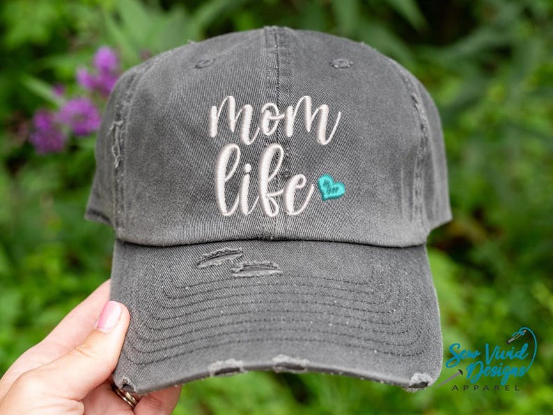 May include: A grey distressed baseball cap with the text "mom life" embroidered in white with a small turquoise heart.