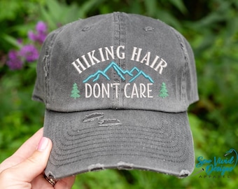 May include: A gray distressed baseball cap with the text "Hiking Hair Don't Care" embroidered in white. The embroidery also includes a blue mountain range and two green pine trees.