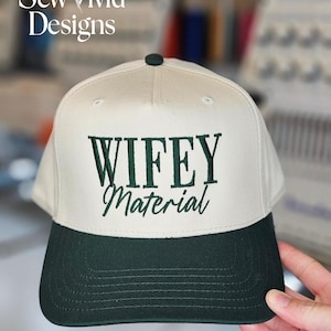 May include: A baseball cap with a cream-colored crown and a dark green brim. The cap features the embroidered text "WIFEY Material" in dark green. The cap has a dark green button on top and the brand name "Sew Vivid Designs" is visible.