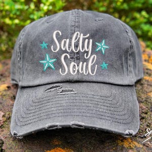May include: A black distressed baseball cap with the text "Salty Soul" in white lettering and teal starfish accents.