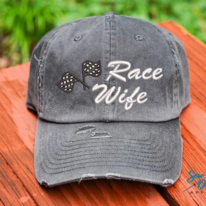 May include: A black distressed baseball cap with a white embroidered design that says "Race Wife" and a checkered flag graphic.