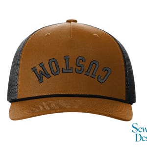May include: A brown trucker hat with a black mesh back and a curved brim. The word "CUSTOM" is embroidered in gray block letters across the front. The hat has a snap closure and a brown button on top.