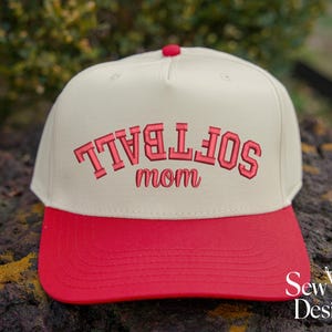 May include: A cream-colored baseball cap with a bright red brim and a red button on top. The front features embroidered red text that reads "SOFTBALL" in an arched block font, with the word "mom" in a cursive script font directly below it.
