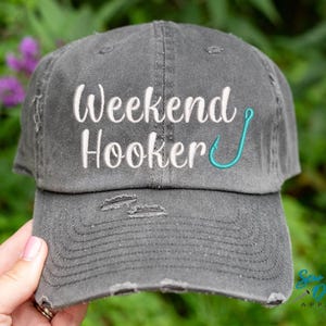 May include: A grey distressed baseball cap with the text "Weekend Hooker" embroidered in white with a turquoise fishing hook.