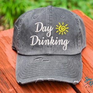 May include: A gray distressed baseball cap with a white embroidered design that says "Day Drinking" with a yellow sun graphic above the text.