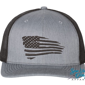 May include: A gray and black trucker hat with a distressed American flag design embroidered on the front. The hat has a black mesh back and a gray front panel.