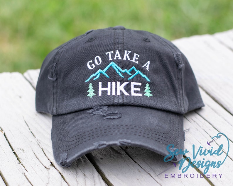 Op de afbeelding: Zwarte, verweerde baseballpet met witte borduurwerk die "Go Take A Hike" zegt met een blauwe bergketen en groene bomen.