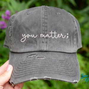 May include: Distressed gray baseball cap with the embroidered phrase "you matter;" in white script. The hat has a pre-worn look with distressed details on the brim and crown. The background is a blurred green with purple flowers.
