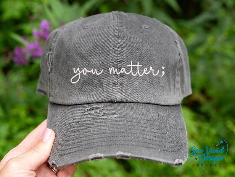 May include: Distressed gray baseball cap with the embroidered phrase "you matter;" in white script. The hat has a pre-worn look with distressed details on the brim and crown. The background is a blurred green with purple flowers.