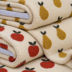 Peut inclure: Un tissu texturé de couleur crème présentant un motif répétitif de pommes rouges et de poires jaune doré. Le matériau possède une surface douce, duveteuse et polaire avec de petites tiges vert foncé sur chaque fruit, disposées en grille.