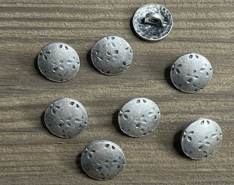 May include: Eight silver-toned buttons with a floral design. The buttons are round and have a raised edge. They are arranged in a circle on a wooden surface.