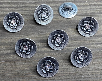 May include: Seven silver-toned buttons with a raised flower design and a single rhinestone in the center. The buttons are arranged in a circle on a wooden surface.