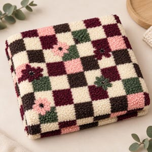 Peut inclure: Une pièce textile touffetée carrée présentant un motif à carreaux dans des tons crème, brun foncé, vert sauge et rose tendre. Certains carrés sont ornés de petits motifs floraux brodés en rose et bordeaux foncé.