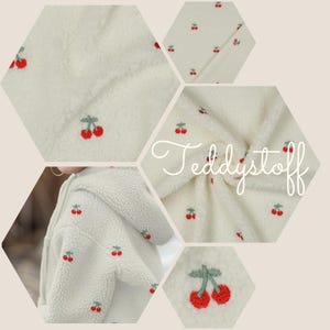 Peut inclure: Tissu sherpa crème avec des cerises rouges et des tiges vertes brodées. L'image montre des gros plans du tissu, un sweat à capuche et le mot "Teddystoff". Le tissu a un aspect doux et texturé, et les cerises sont espacées uniformément.