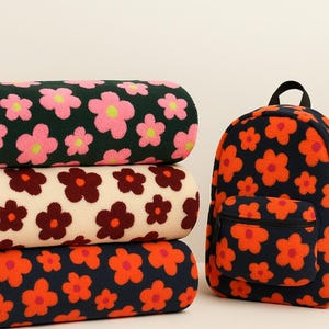 Könnte beinhalten: Drei gerollte Decken und ein Rucksack, alle mit einem sich wiederholenden Blumenmuster. Die Decken zeigen Blumen in Dunkelgrün mit rosa Akzenten, Creme mit Kastanienbraun und Marineblau mit Orange. Der Rucksack hat eine marineblaue Basis mit orangefarbenen Blumen.