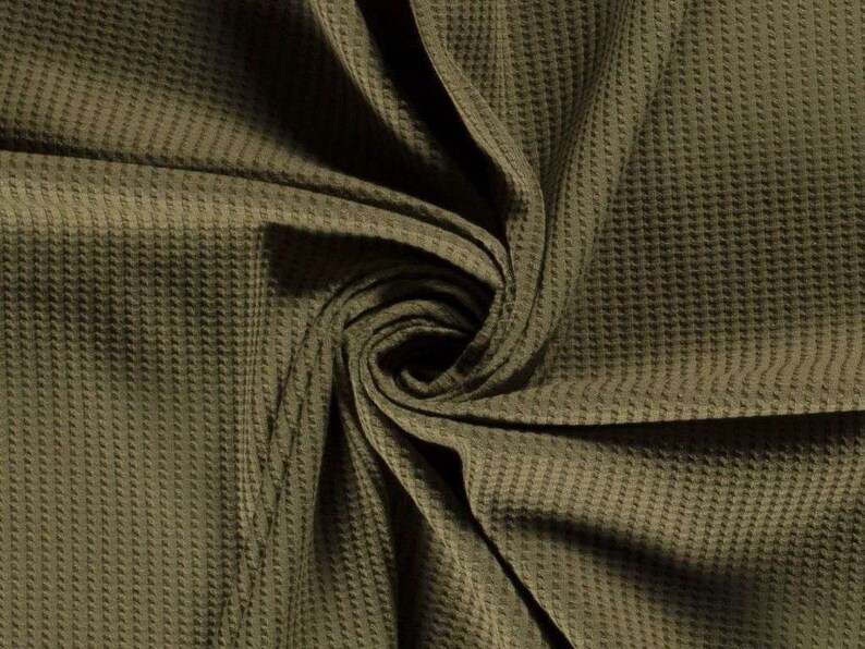 May include: Close-up of a textured, olive green waffle knit fabric. The fabric is folded and twisted, showing the texture and drape of the material.