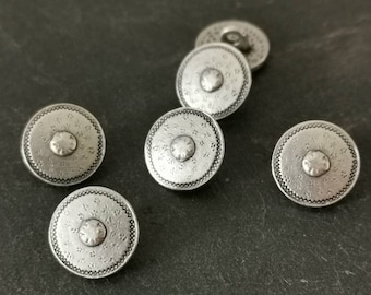 May include: Six silver-toned metal buttons with a decorative design. The buttons have a raised center and a circular pattern around the edge.