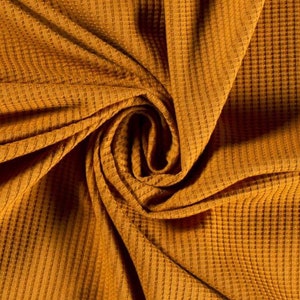 May include: A close-up of a textured, mustard yellow waffle knit fabric. The fabric is folded and twisted, creating a swirling pattern.