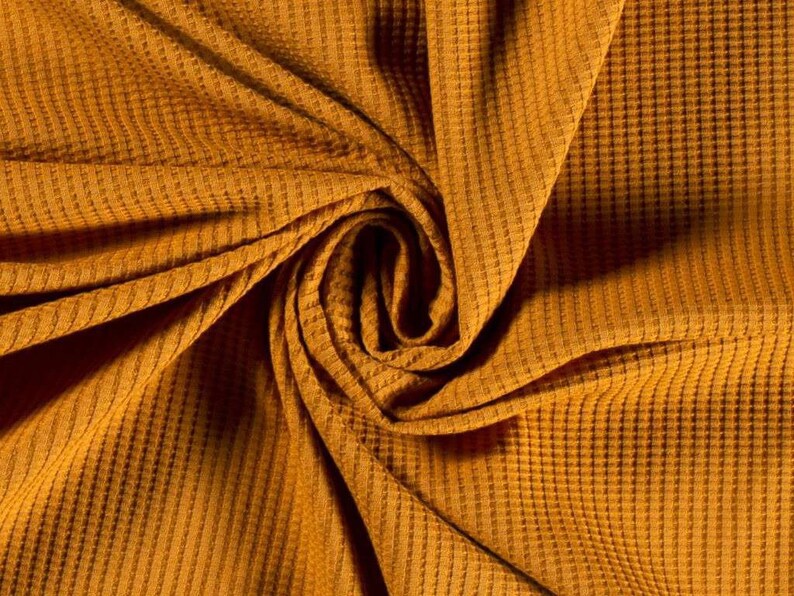 May include: A close-up of a textured, mustard yellow waffle knit fabric. The fabric is folded and twisted, creating a swirling pattern.