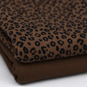 Könnte beinhalten: Ein gefaltetes Stück gerippter Stoff mit brauner Grundfarbe und schwarzem Leopardenmuster. Der Stoff liegt auf einem einfarbigen, dunkelbraunen Rippenstoff, wodurch die Textur und der Kontrast zwischen den beiden Materialien deutlich werden.