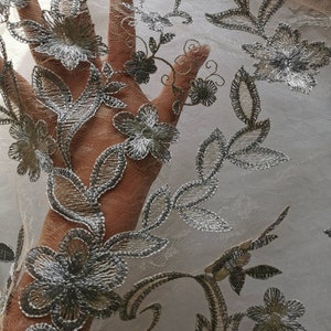 May include: A close-up of a sheer fabric with a silver floral embroidery pattern. The fabric is delicate and has a soft, flowing texture.