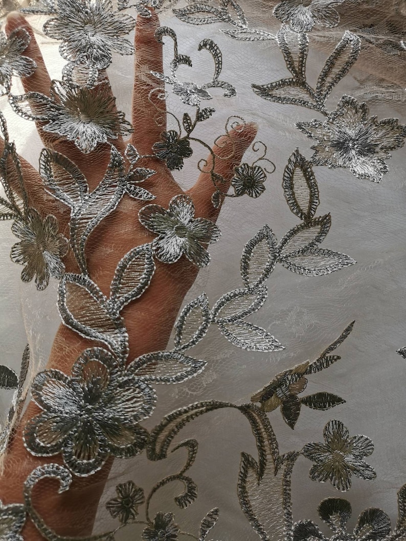 May include: A close-up of a sheer fabric with a silver floral embroidery pattern. The fabric is delicate and has a soft, flowing texture.