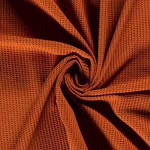 May include: Close-up of a rust-colored waffle knit fabric. The fabric is folded and twisted, creating a swirling pattern.