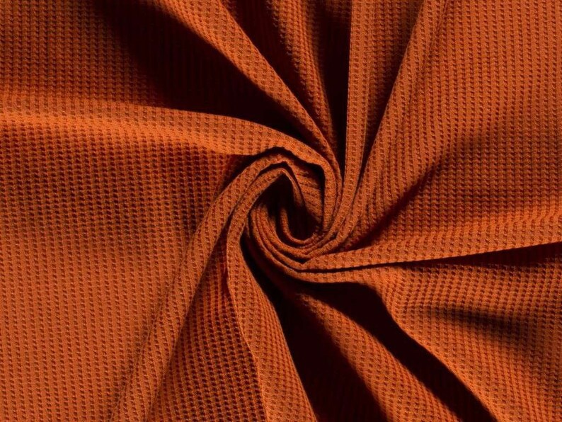 May include: Close-up of a rust-colored waffle knit fabric. The fabric is folded and twisted, creating a swirling pattern.