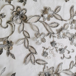 May include: Close-up of delicate silver and white floral lace fabric. The lace features a repeating pattern of flowers and leaves.