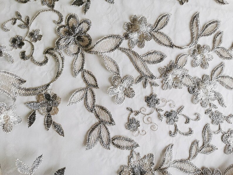 May include: Close-up of delicate silver and white floral lace fabric. The lace features a repeating pattern of flowers and leaves.