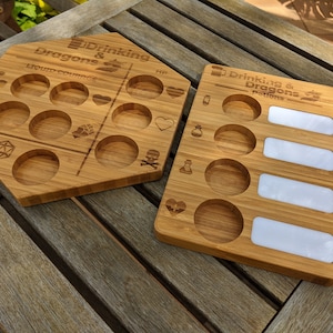 May include: Two wooden game boards for a Drinking and Dragons game. The first board has 10 round indentations for drinks and is shaped like a hexagon. The second board has 5 round indentations for drinks and 4 rectangular spaces for writing. Both boards have the text "Drinking & Dragons" on them.