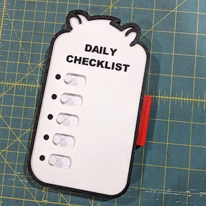 May include: A white daily checklist with a black outline and a red side grip. The checklist has the words "DAILY CHECKLIST" printed in black and five toggle switches. The checklist is on a green surface.