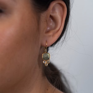 May include: Gold-tone earrings with a square, iridescent green gemstone and three smaller, round, clear gemstones. The earrings are hanging from a person's ear.